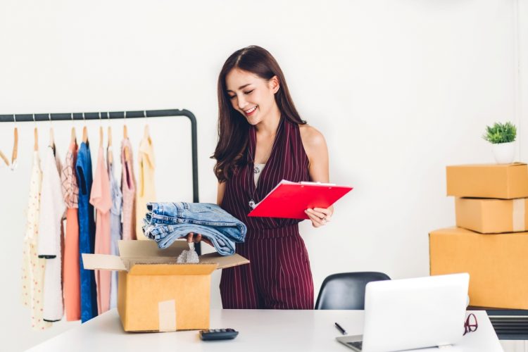 Young woman freelancer working sme business online shopping and packing clothes with cardboard box at home - Business online shipping and delivery concept